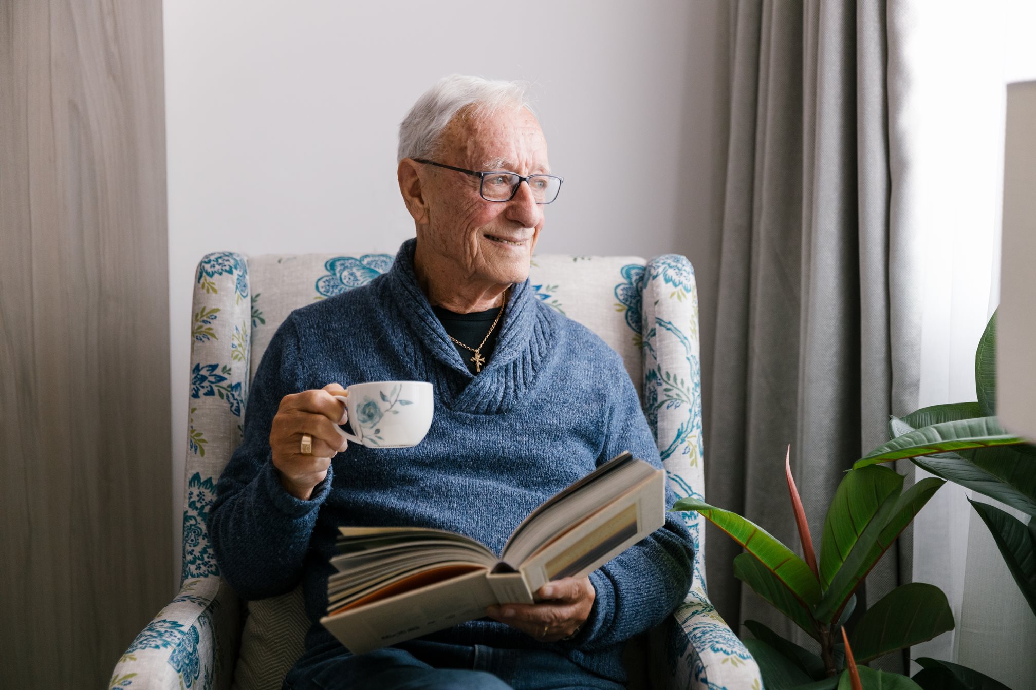 an aged care resident reading a book in his bedroom with a tea in his hand